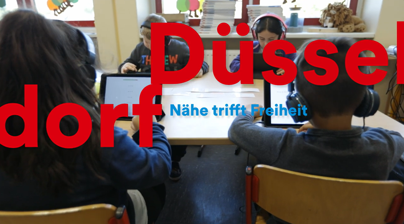 Schülerinnen und Schüler sitzen an einem Gruppentisch und arbeiten mit Tablets sowie Kopfhörern im Unterricht. Über dem Bild liegt der Schriftzug „Düsseldorf – Nähe trifft Freiheit“.
