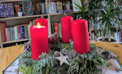 Foto von einem Adventskranz mit roten Kerzen, von denen eine brennt, vor einem Bücherregal.