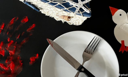 Fotografie mit einem weißen Teller, Besteck, einem Fischernetz, einer Möwe aus Papier und einem blutroten Handabdruck
