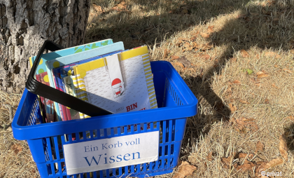Foto von einem blauen Tragekorb mit der Aufschrift "Ein Korb voll Wissen" der auf einer Wiese an einem Baum steht. In dem Korb sind Kinderbücher.