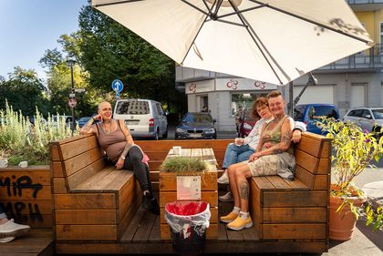 Auf einer hufeisigenförmigen Holzbank befinden sich sitzen drei weibliche Personen mit kurzen Haaren. 