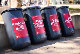 Vier Abfallbehälter mit rot-weißen Motivaufklebern an Mauer gelehnt