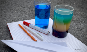 Foto mit weißem, karierten Papier, Stiften und zwei Gläsern mit bunten Flüssigkeiten darin