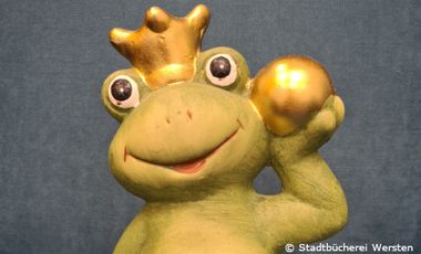 Froschfigur mit Krone und goldenem Ball aus Keramik vor grauem Hintergrund.