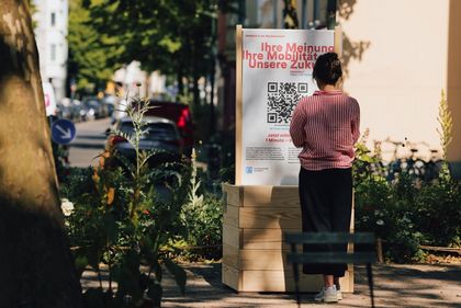 Person steht vor einer Stele auf der Text und ein QR-Code abgebildet sind.