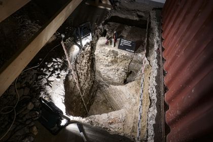 Es handelt sich um ein Foto der Fundstelle der ehemaligen Festungsmauern unter dem Behrensbau. Das Foto ist von oben aufgenommen worden und man blickt in eine rechteckige Grube. Dort sind rote Ziegel in mehreren Lagen zu erkennen, deren Position zueinander darauf schließen lässt, dass hier zwei Mauern der ehemaligen oberrheinischen Bastion im rechten Winkel aufeinander treffen. Auf dem Foto sind außerdem noch Scheinwerfer und Messwerkzeuge zu sehen. Die Zollstöcke zeigen an, dass die Grube etwa 2 Meter 50 mal 1 Meter 50 groß und etwa 1 Meter 60 tief ist.
