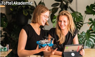 Foto mit zwei jungen Frauen, die an einem selbst gebautem Roboter mit einem Tablet tüfteln. Schriftzug am oberen Rand des Fotos: Tüftelakademie.