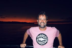 Portraitaufnahme von Rodrigo Butori in rosafarbenem T-Shirt, auf dem mittig das PlasticFisherman-Logo aufgedruckt ist. Im Hintergrund ist der Sonnenuntergang über dem Meer zu sehen. 