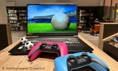 Aufgeklappter Laptop mit einem Bild auf dem Monitor, auf dem ein Fußball auf einem Rasen zu sehen ist. Dazu Gamecontroller vor dem Laptop und kleine Fußbälle.