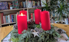 Foto von einem Adventskranz mit roten Kerzen, von denen eine brennt, vor einem Bücherregal.