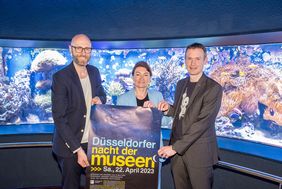 Miriam Koch, Tobias Etzel und Dr. Jochen Reiter präsentieren das Poster zur Nacht der Museen vor dem Korallen-Riffbecken im Aquazoo
