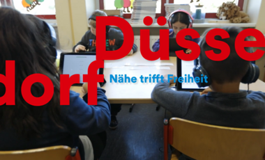 Schülerinnen und Schüler sitzen an einem Gruppentisch und arbeiten mit Tablets sowie Kopfhörern im Unterricht. Über dem Bild liegt der Schriftzug „Düsseldorf – Nähe trifft Freiheit“.