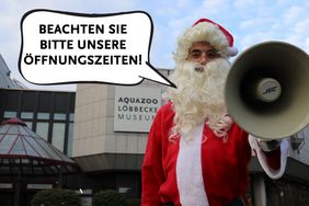 Ein Weihnachtsmann steht mit einem Megafon in der linken hand vor dem Aquazoo Löbbecke Museum. In einer Sprechblase ist folgender Text aufgeführt: "Beachten Sie bitte unsere Öffnungszeiten!"