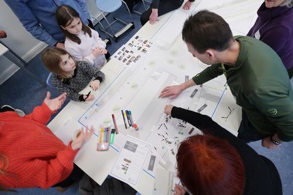Aufblick auf einen planerischen Entwurf an einem großen Tisch; auf dem Entwurf befinden sich farbige Stifte. In einer Runde um den Tisch diskutieren Kinder und Erwachsene über dem Entwurf. 