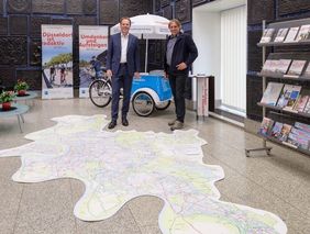 Foto von Oberbürgermeister Dr. Stephan Keller (l.) und Mobilitätsdezernent Jochen Kral im Foyer des Rathauses. Vor ihnen der Düsseldorfer FAhrradnetzplan als Vinylfototeppich.