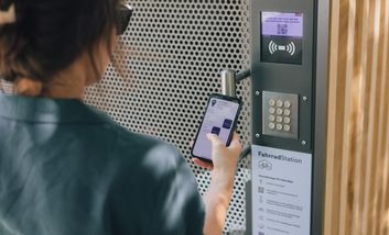 Frau mit einer Sonnenbrille steht vor dem Terminal einer Fahrradstation. Sie hält ein Handy. Displays vom Handy und Terminal sind sichtbar.