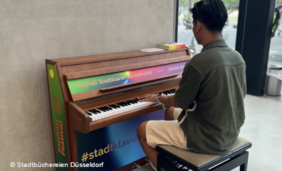 Foto von einem jungen Mann, der Klavier spielt. Das Klavier steht in der Glashalle im Eingang der Zentralbibliothek im KAP1