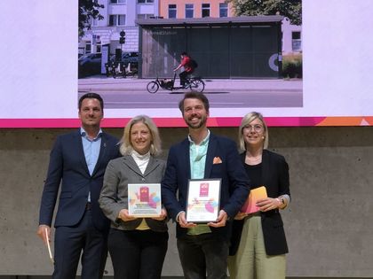Zwei Männer und zwei Frauen stehen frontal in einer Reihe und lächeln. In der Mitte halten eine Frau und ein Mann jeweils eine Urkunde zur Auszeichnung der Mobilitäts- und Fahrradstationen beim Eurobike Award.