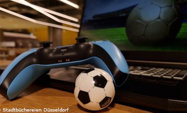 Aufgeklappter Laptop mit einem Bild auf dem Monitor, auf dem ein Fußball auf einem Rasen zu sehen ist. Dazu ein Gamecontroller vor dem Laptop und ein kleiner Fußball.
