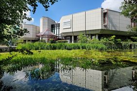 Das Gebäude des Aquazoo von außen. Davor ist ein mit Pflanzen bewachsener Teich zu sehen. 