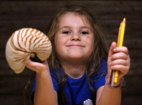 Ein Mädchen in blauem T-SHirt hält inseiner linken Hand einen Bleistift und in seiner rechten Hand die Schale eines Nautilus (Kopffüßers).