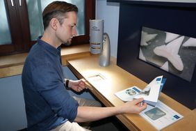 Ein Mann in einem blauen Hemd bedient ein Videomikroskop und blättert dabei in einem Bestimmungsbuch. Auf einem Bildschirm sind Haizähne zu sehen. 