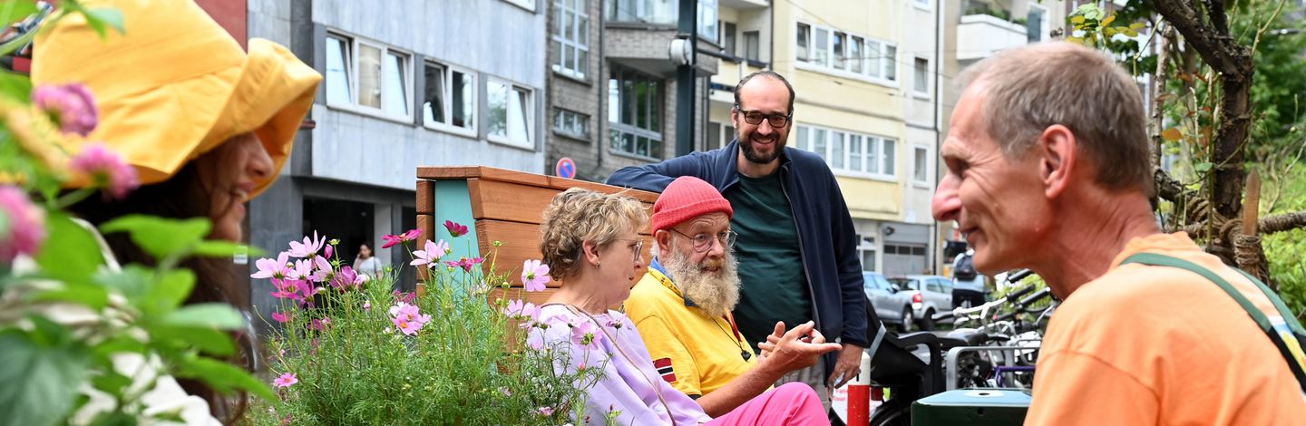 Zwei Personen sitzen auf hochwertigen hölzernen Sitzmöglichkeiten, umgeben von blühenden Hochbeeten. Sie sind in einem Gespräch vertieft. Im Vordergrund sitzt eine weitere Person.