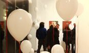 Menschen betrachten Bilder an einer weißen Wand, im Vordergrund schweben weiße Luftballons