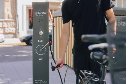 Person hält an einer Reparaturstation für Fahrräder eine Luftpumpe. Vorne steht ein Fahrrad.