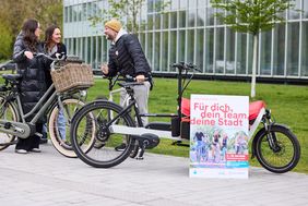 Drei Personen stehen mit ihren Rädern beieinander und unterhalten sich. An ein Lastenrad ist rechts im Bild das STADTRADELN-Plakat angelehnt, Motto "Für dich, dein Team, deine Stadt"