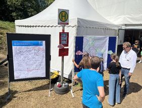 Zu sehen ist eine Haltestelle mit einem Zusatzschild "Mitmach-Haltestelle", Darum Kinder und Mitarbeiter der Verwaltung, sowie Pinnwände auf denen die Kinder und Jugendliche  Kommentare schreiben.