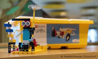 Im Vordergrund steht ein zusammengebauter Lego-Spike Roboter. Im Hintergrund eine Legokiste