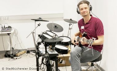 Foto mit Mann der vor digitalem Schlagzeug mit eBass sitzt.