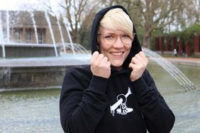 Eine blonde Frau steht vor einem Brunnen im Nordpark und trägt den schwarzen Hoodie mit dem Motiv "Tentakel greift Rheintum". Sie hält mit ihren Händen die Kapuze auf dem Kopf fest.r