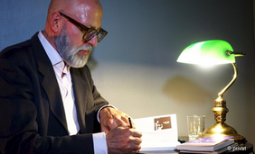 Ein Mann sitzt links im Bild, er trägt Anzug und eine schwarze Hornbrille und Bart. Er schreibt etwas an einem Schreibtisch. Auf dem Tisch steht eine kleine grüne Lampe.