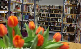 Foto von Bücherregalen in der Bücherei Eller mit einem orangen Tulpenstrauß im Vordergrund.