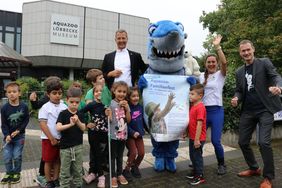 Josef Hinkel (Bürgermeister Düsseldorf), Silke Heise (Weltretterbande) und Dr. Jochen Reiter (Aquazoo-Direktor) inmitten einer Kindergruppe vor dem Gebäude des Aquazoo Löbbecke Museum. Ein Hai (Maskottchen) hält das Poster zum Aquazoo-Familienfest in den Flossen.