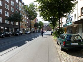 Foto von Bilker Allee und einem Radfahrer der auf der rechten Fahrspur an parkendem Auto vorbei fährt.
