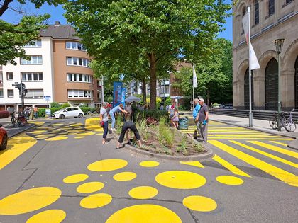 Nachbarschaftszone auf dem Paulusplatz; auffällige, farbige Fahrbahnmarkierungen unterschiedlicher Größen – Punkte, Striche und Linien – befinden sich um eine Verkehrsinsel mit einem Baum und Hochbeet. Eine 