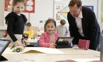 Zwei Schülerinnen zeigen Stadtdirektor Burkhard Hintzsche in einem Klassenraum einen kleinen Roboter. Im Hintergrund arbeiten weitere Kinder an digitalen Lernprojekten.