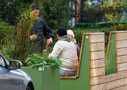 Zwei Personen sitzen auf hochwertigen hölzernen Sitzmöglichkeiten einer Nachbarschaftszone, umgeben von einem Blumentopf  mit Gräsern. 