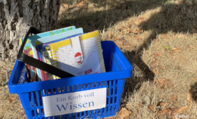 Foto von einem blauen Tragekorb mit der Aufschrift "Ein Korb voll Wissen" der auf einer Wiese an einem Baum steht. In dem Korb sind Kinderbücher.