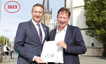 Stadtdirektor Burkhard Hintzsche (r.) erhält von Thomas Schürmann, Regierungspräsident der Bezirksregierung Düsseldorf, einen Förderbescheid. Beide blicken in die Kamera.