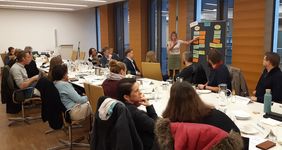 Viele Personen an langem Tisch, Vortragende an Pinwand