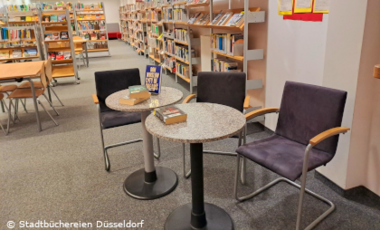 Foto aus einer Bibliothek mit Bücherregalen im Hintergrund und zwei runden Tischen mit Stühlen im Vordergrund. Auf den Tischen liegen Bücher.