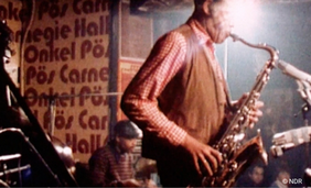 Ein Saxophonist steht in der Mitte des Bildes. Im Hintergrund eine sitzende Person mit Schiebermütze.