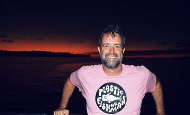 Rodrigo Butori steht vor einem Sonnenuntergang am Meer und schaut in die Kamera. Erträgt ein rosafarbenes T-Shirt, auf dem das Logo der PlasticFisherman-Bewegung abgebildet ist.