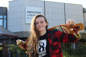 Eine Frau mit langen, braunen, gelockten Haaren steht vor dem Aquazoo Löbbecke Museum und  trägt ein schwarzes T-Shirt mit Tenntakel-Motiv. Darüber trägt sie geöffnet eine rot-karrierte Jacke und ein Longboard über den Schultern.